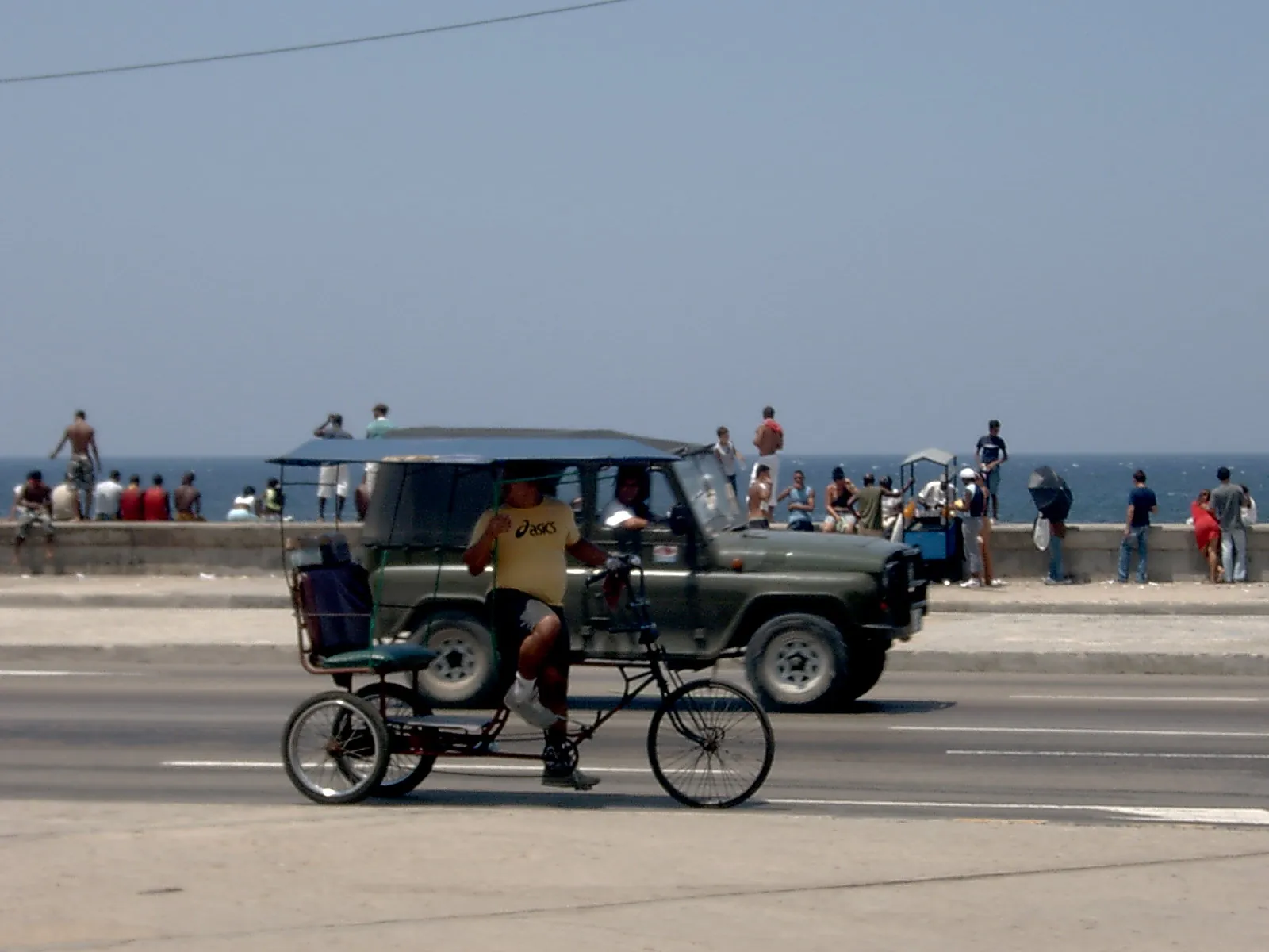 Un bicitaxi y un jeep comparten la avenida del Malecón mientras la gente disfruta del muro